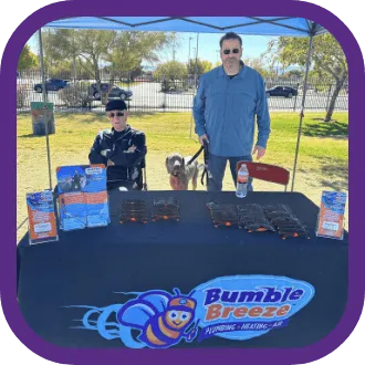 Two people and a dog are at an outdoor event booth for Bumble Breeze, a heating and air company. The table displays brochures, sunglasses, and an "about us" section, with trees and a fence in the background.