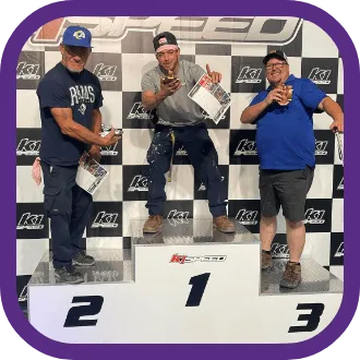 Three smiling men stand on a winners’ podium marked 1, 2, and 3 at K1 Speed, holding certificates and cans, celebrating their achievements. The about us section comes alive against a black and white checkered wall in the background.