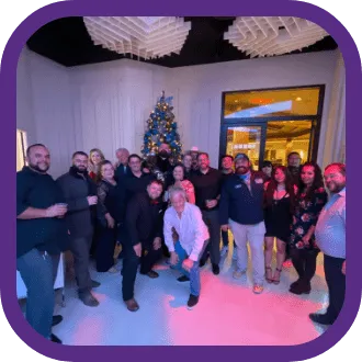 A group of people pose and smile in front of a decorated Christmas tree indoors, with festive lighting and modern decor visible in the background—a perfect moment that captures the spirit of togetherness seen on our About Us page.