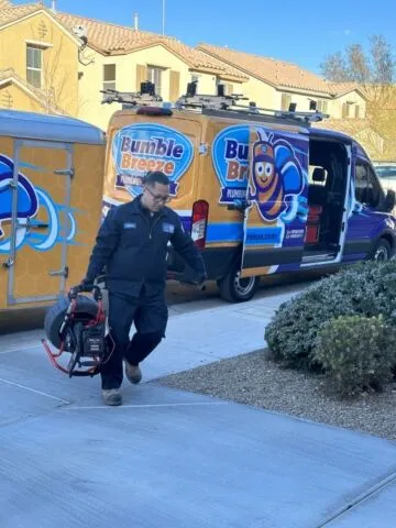 A worker in uniform carries equipment in front of a Bumble Breeze service van, which is brightly decorated with a cartoon bee logo, parked on a residential street near tan houses and bushes.