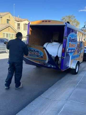 A person stands next to a Bumble Breeze service trailer parked on a residential street, unloading or loading equipment from the open back door.