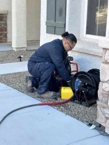 A person in work overalls kneels on gravel outside a house, operating a large hose reel and yellow tool near a window.