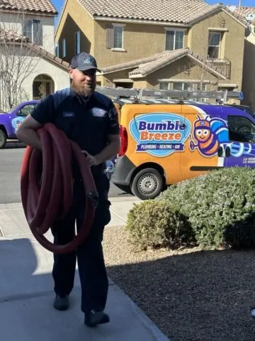 A man in a work uniform carries red hoses outside a house, with a Bumble Breeze plumbing and HVAC van featuring a cartoon bee parked in the driveway.
