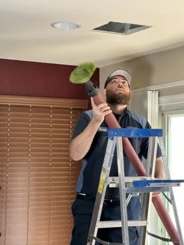A man standing on a ladder uses a vacuum with a brush attachment to clean an air vent in the ceiling of a room with red walls and closed blinds.