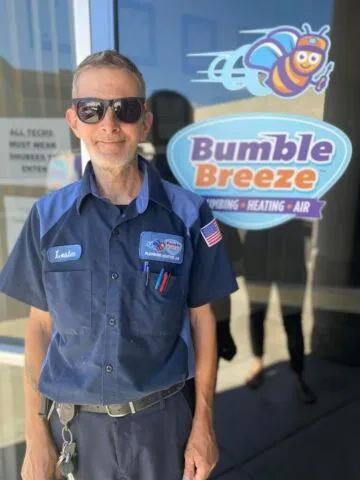 A man in a blue work uniform and sunglasses stands in front of a glass door with a "Bumble Breeze Plumbing Heating Air" sign featuring a cartoon bee logo.