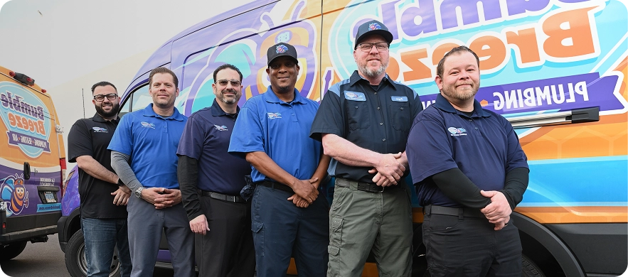 Six men in work uniforms stand smiling in front of a colorful van with "Plumbing" visible on the side, suggesting a plumbing or repair service team.