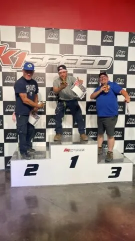 Three men stand on a winners' podium at an indoor kart racing venue, holding trophies and certificates. The man in the center (first place) cheers, while the others stand on second and third place podiums. Racing logo backdrop behind them.