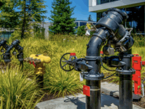 %Imagetitle% | Bumble Breeze Plumbing, Heating &Amp; Air Outdoor view of large black water pipes with valve wheels, red tags, and a yellow hydrant surrounded by tall green grass and trees near a modern building.