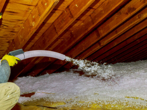 A person in yellow gloves is using a hose to blow loose fill insulation into an attic, filling the floor space between wooden beams.
