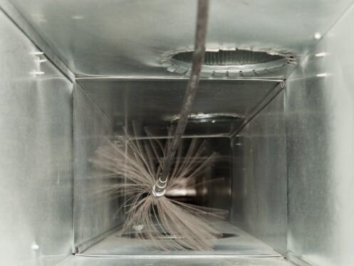 A close-up view inside a metal air duct, showing a rotating brush cleaning tool attached to a flexible rod used for duct cleaning.