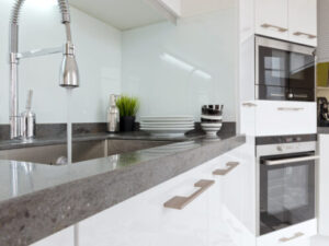 %Focuskw% | Bumble Breeze Plumbing, Heating &Amp; Air Modern kitchen with a gray countertop, stainless steel sink and faucet, stacked white dishes, bowls, a plant, and built-in appliances including an oven and microwave in glossy white cabinets.