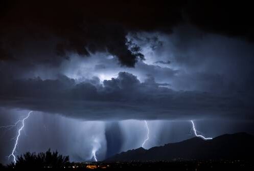 Lightning streaks across a dark night sky beneath thick storm clouds, illuminating rain falling over distant mountains and a barely visible landscape below.