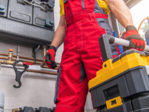 A plumber wearing red overalls and gloves stands in a utility room, holding a toolbox with plumbing pipes, valves, and equipment visible in the background.