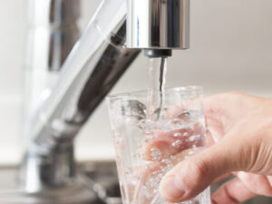 A hand holds a clear glass under a kitchen faucet as water pours in, filling the glass with fresh water and creating bubbles.