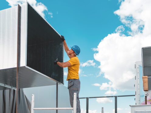 A worker wearing a blue hard hat, yellow shirt, and black gloves installs or inspects a large metal HVAC duct outdoors under a bright blue sky with scattered clouds.
