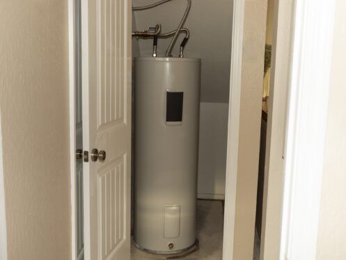 A water heater is installed in a small utility closet with white walls and a partially open door. Pipes are connected at the top of the unit.