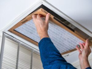 %Focuskw% | Bumble Breeze Plumbing, Heating &Amp; Air A person’s hands are installing or replacing a rectangular air filter in a ceiling vent. The person is wearing a blue long-sleeve shirt and the vent cover is open, revealing the filter slot.