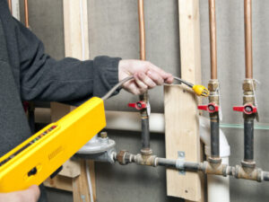 A person uses a yellow handheld gas leak detector to check for leaks in copper pipes with red-handled valves mounted on a wall frame.