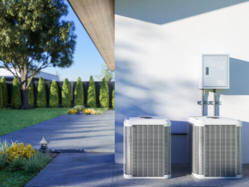 Two outdoor air conditioning units are installed next to a modern house, with a green lawn, trees, and hedges visible in the background on a sunny day.