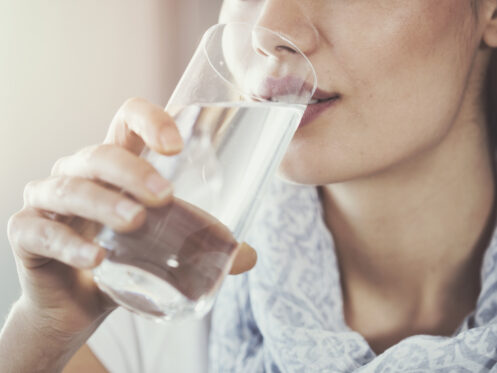 A woman wearing a patterned scarf drinks a glass of water, holding the glass close to her mouth with her right hand. The image focuses on her face and hand.