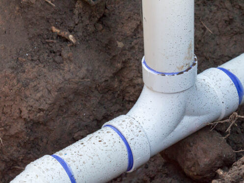 White PVC pipes with blue seals are joined at a T-junction and partially buried underground, surrounded by soil and dirt.