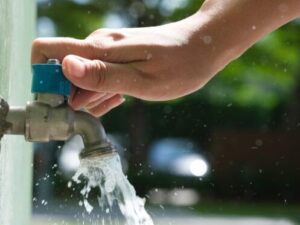 %Focuskw% | Bumble Breeze Plumbing, Heating &Amp; Air A hand turns on an outdoor metal water tap, with water flowing out. The background is blurred with greenery and sunlight.
