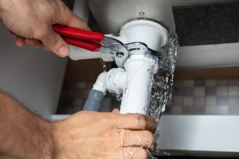 A person uses a wrench to fix a leaking pipe under a sink, with water spraying out from the pipe joint.