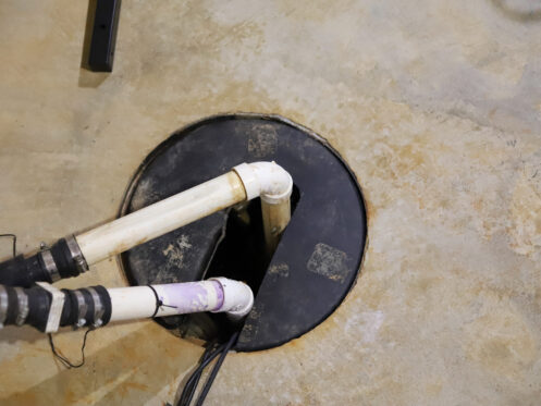 Two white PVC pipes and a black cable enter a round black sump pump cover set in a concrete floor, showing a typical basement sump pump setup.