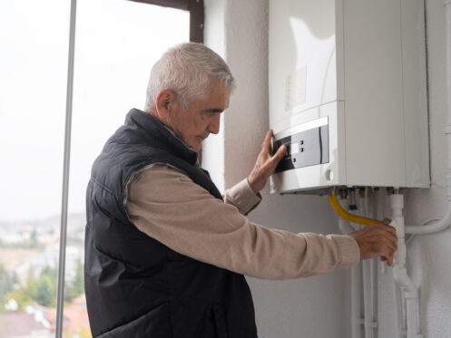 An older man wearing a black vest adjusts controls on a white wall-mounted gas boiler inside a bright room, focusing on the device with one hand on the control panel and the other on a pipe.