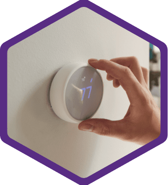 A hand adjusts a modern digital thermostat mounted on a wall, displaying the temperature set to 77 degrees.
