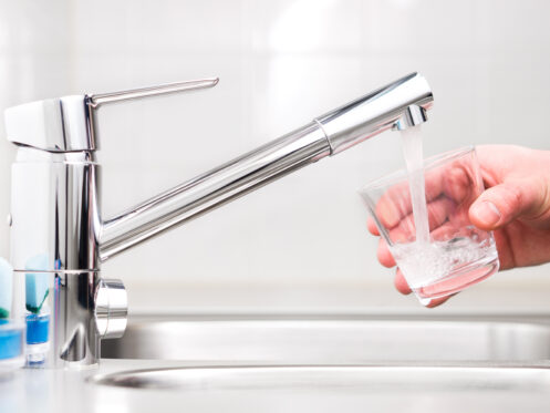 A hand holds a glass under a modern kitchen faucet as water flows into the glass. The sink and faucet are metallic and the background is out of focus, showing a clean tiled wall.
