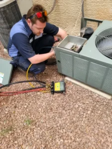 A technician in a blue uniform kneels on gravel while servicing an outdoor air conditioning unit with tools and diagnostic equipment connected to the unit.