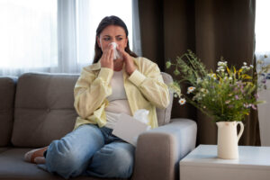 A woman sits on a couch, holding a tissue to her nose as if sneezing or wiping it. She looks unwell. A box of tissues sits beside her, and a vase of flowers is on a table in the foreground.