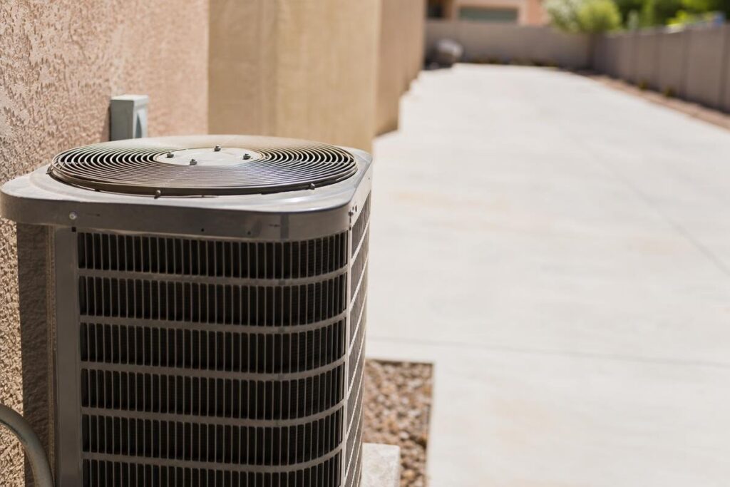 An outdoor air conditioning unit sits next to a beige stucco wall on a concrete pad, with a long, empty driveway stretching into the background on a sunny day.