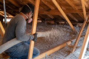 A person wearing a mask and gloves is spraying loose insulation material in an attic space, filling the floor between wooden beams.