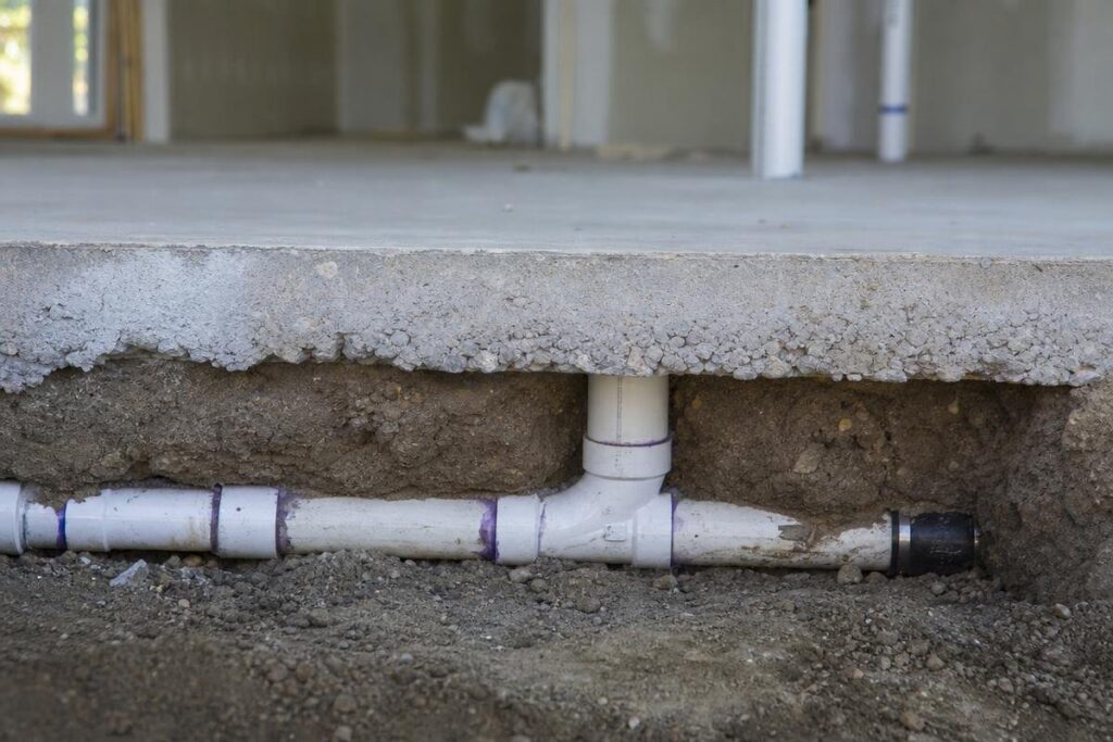 A section of concrete floor is cut away, revealing white PVC plumbing pipes beneath the surface embedded in soil, with one vertical pipe connecting to horizontal pipes.