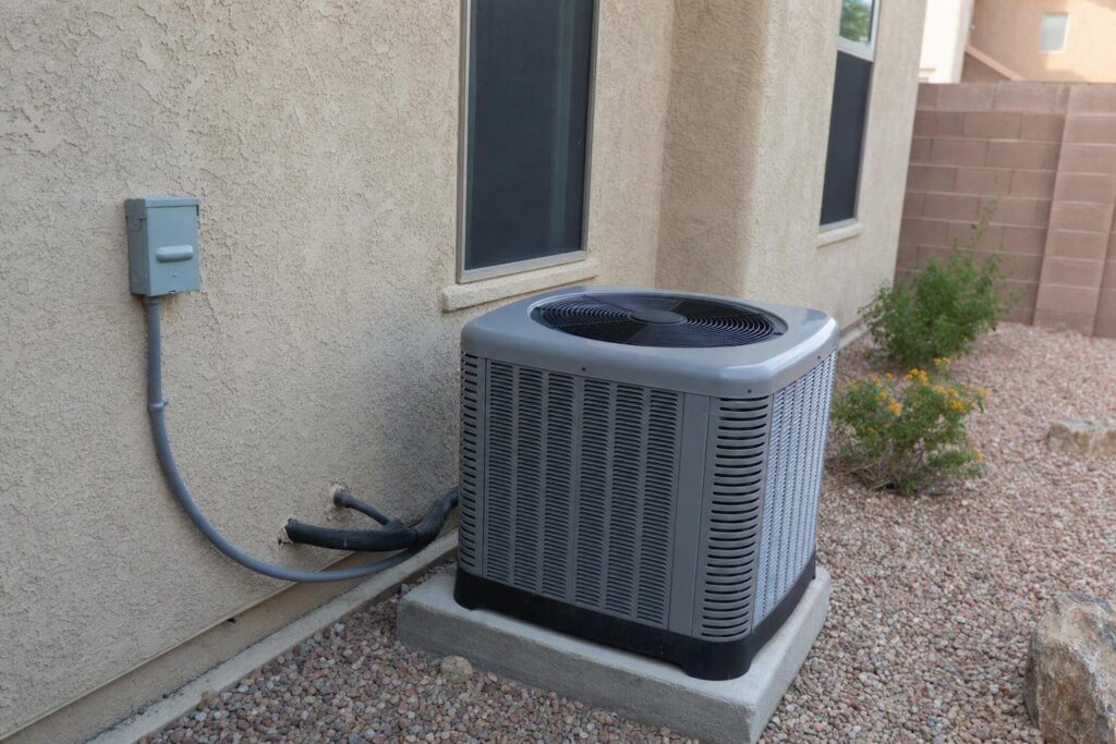 A central air conditioning unit sits on a concrete pad next to a beige stucco house with gravel landscaping and small bushes visible nearby.