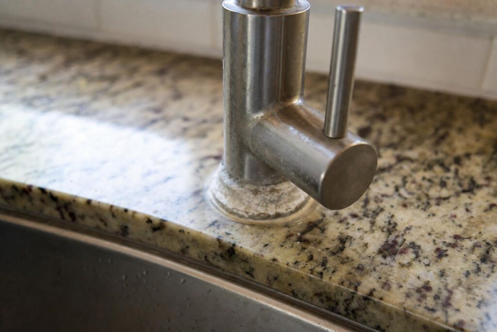 Stainless steel faucet on a granite countertop with visible white hard water stains and mineral buildup at the base near a kitchen sink.