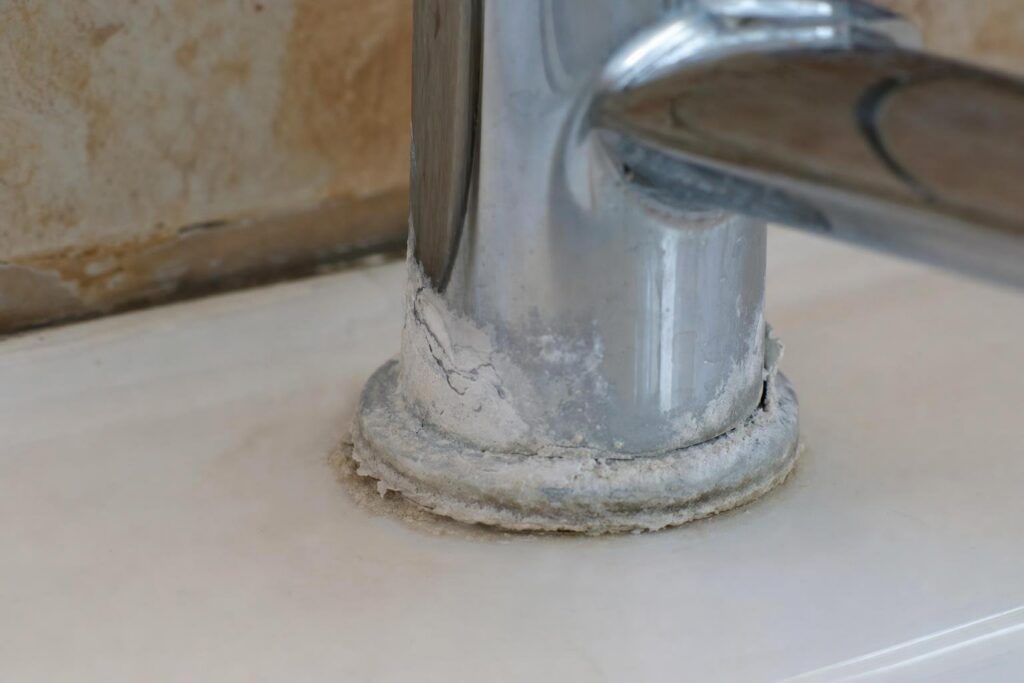 Close-up of a chrome faucet base with noticeable white limescale buildup and mineral deposits on the sink surface and around the bottom of the faucet.