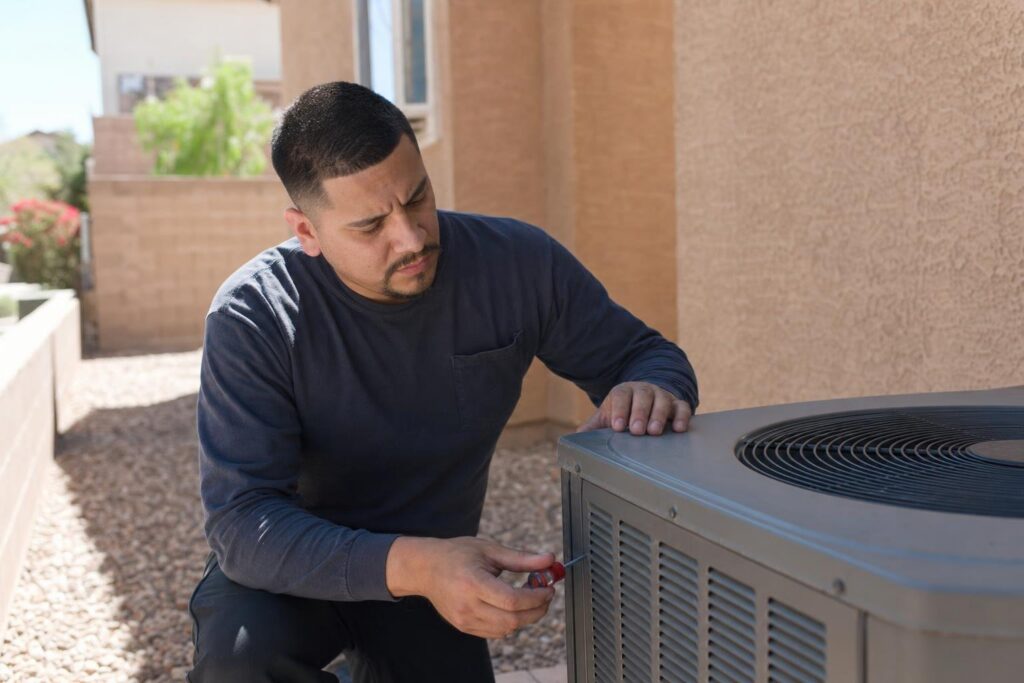 A man in a dark shirt uses a tool to work on an outdoor air conditioning unit next to a house.