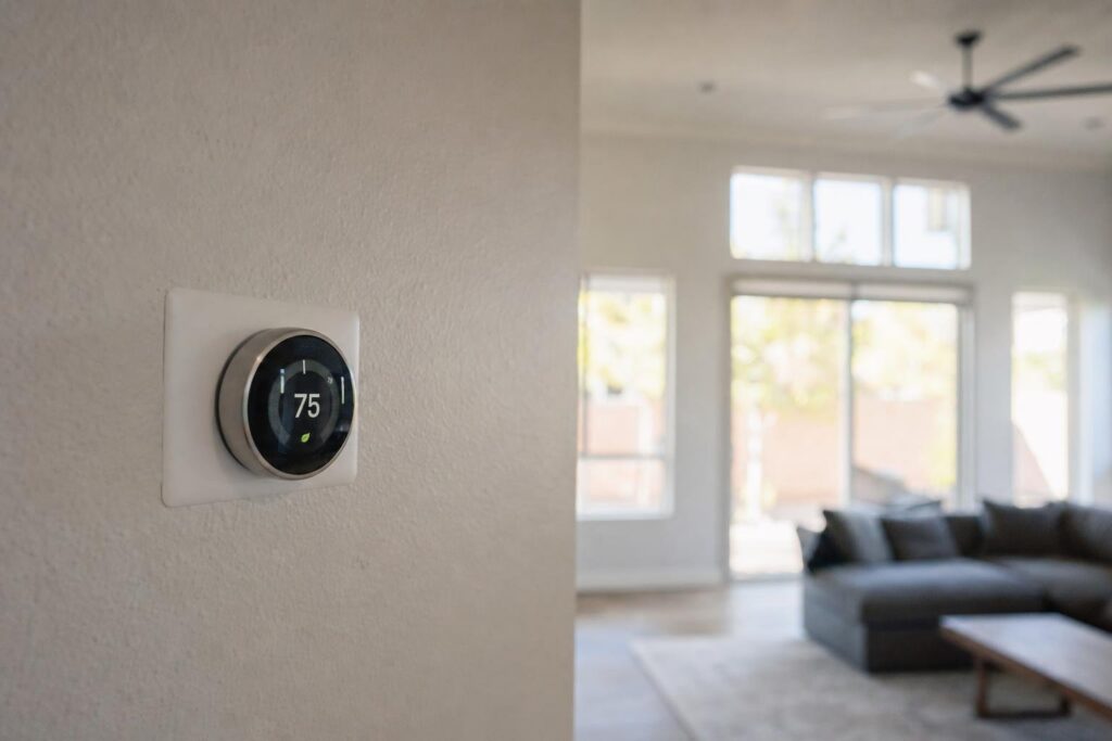 A smart thermostat set to 75 degrees is mounted on a wall in a modern, bright living room with large windows, a ceiling fan, and a couch.