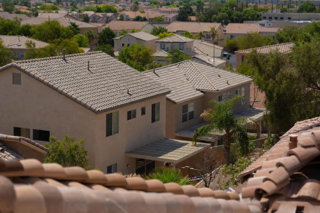 Suburban neighborhood with beige stucco houses and tiled roofs, surrounded by trees and greenery. The photo is taken from an elevated position, showing multiple homes in a residential area under bright daylight.