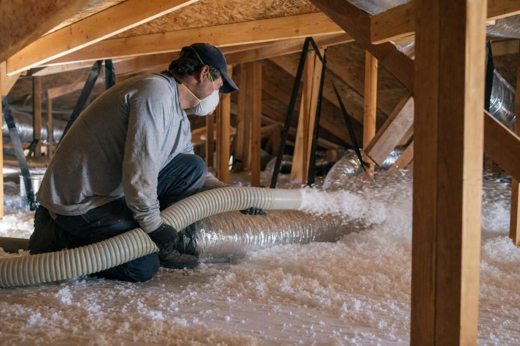 A person wearing a mask and cap kneels in an attic, blowing loose-fill insulation onto the floor with a large hose. Exposed beams and ductwork are visible.