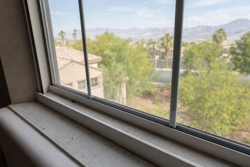 A window with a white frame looks out onto a suburban neighborhood with trees, houses, and distant mountains under a partly cloudy sky.