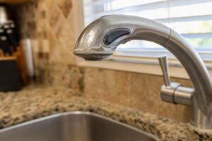 A kitchen sink with a stainless steel faucet showing a buildup of white mineral deposits near the spout, set in a granite countertop with a window and knife block in the background.