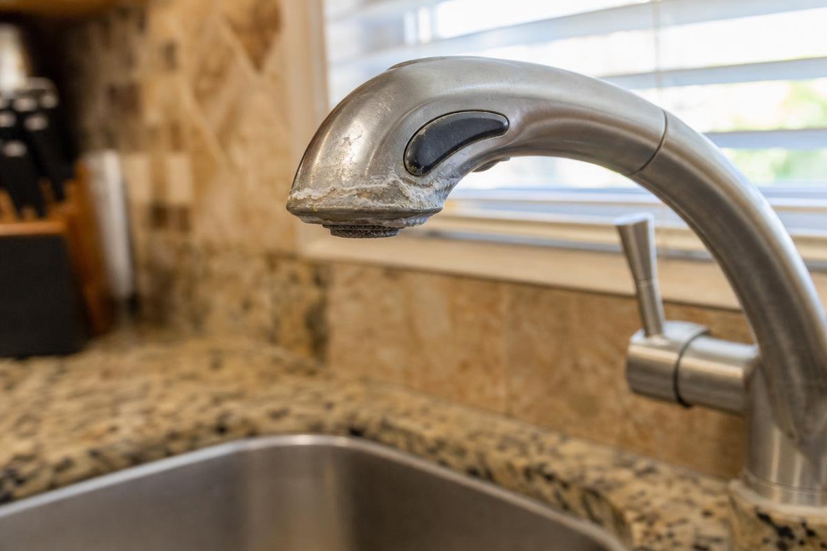 A kitchen sink with a stainless steel faucet showing a buildup of white mineral deposits near the spout, set in a granite countertop with a window and knife block in the background.