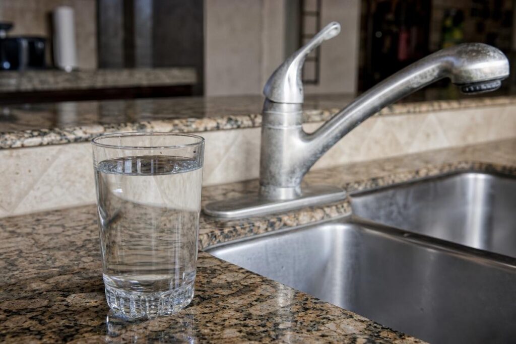 A glass of clear water sits on a granite kitchen countertop next to a stainless steel sink and faucet.