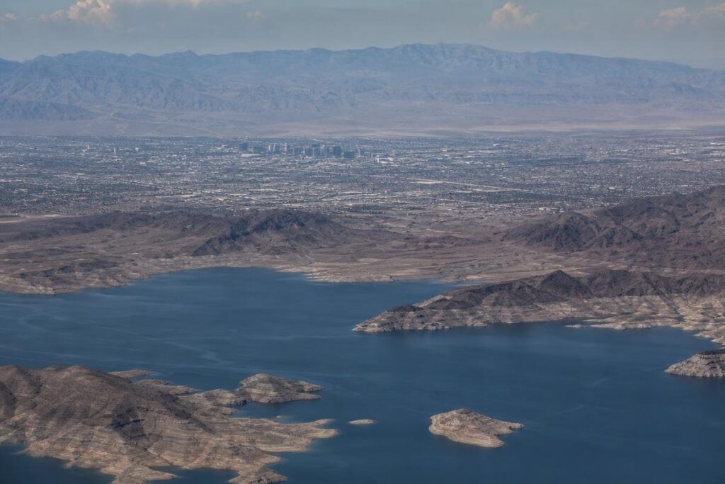 Aerial view of a large blue lake surrounded by dry, rocky hills with a distant city skyline and mountains in the background under a hazy sky.
