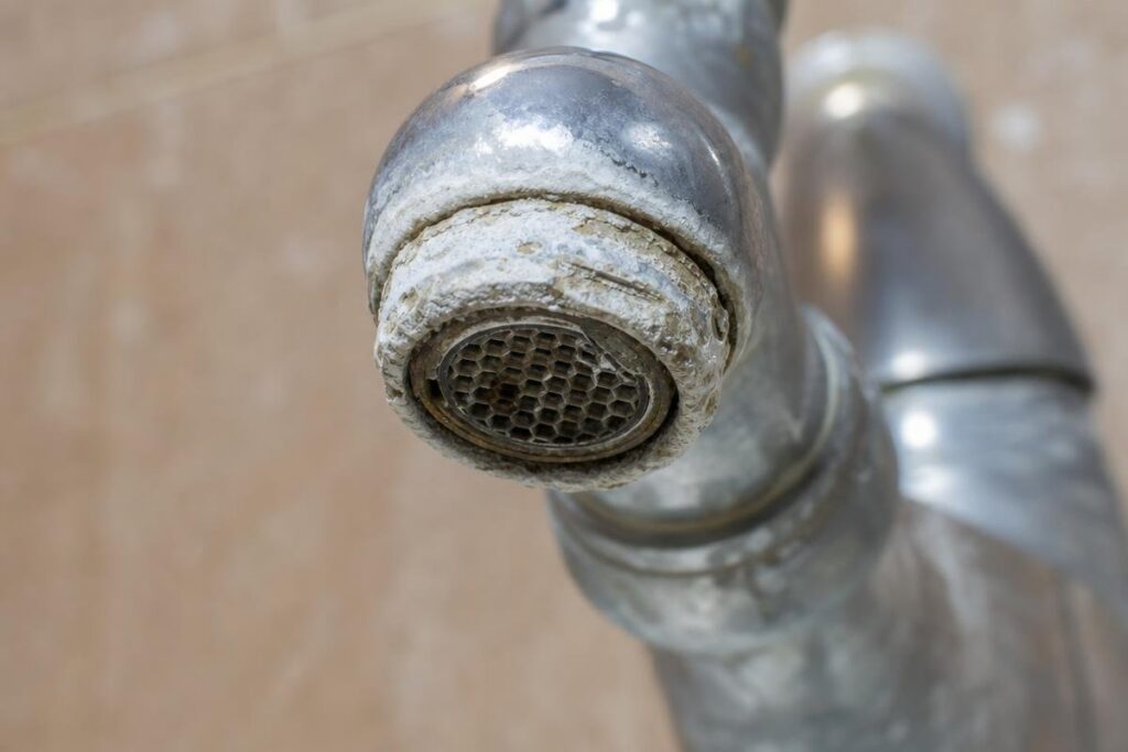 Close-up of a metal faucet with visible white limescale buildup and mineral deposits on the spout, indicating hard water issues. Background is out of focus.