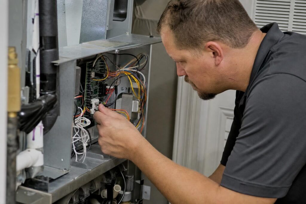 A man in a gray shirt repairs or inspects wiring in an open electrical panel, focusing on connections and components inside a metallic unit.